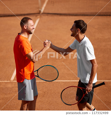 Two smiling men, tennis players on clay court exchanging handshake before game session, showing teamwork and readiness for good training Two smiling men, tennis players on clay court exchanging handshake before game session, showing teamwork and readiness for good training 117332499