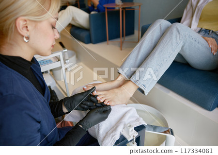 Female pedicure specialist filing down toe nail plate with polishing brush 117334081