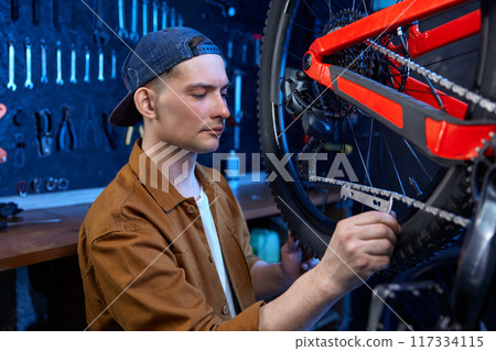 Young repairman providing bike chain stretch measurement at workshop 117334115