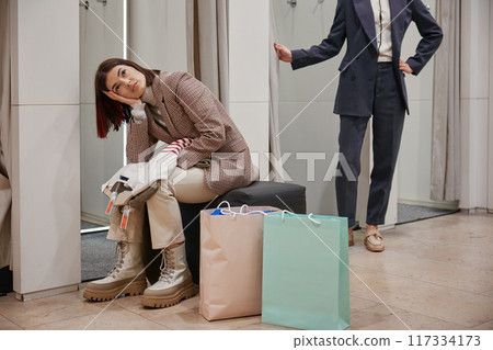 Bored female feeling tired while her friend fitting new clothes attire Bored female feeling tired while her friend fitting new clothes attire 117334173