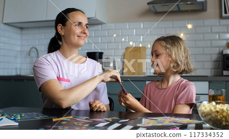 Playing a board game in the kitchen, cheerful young mother and her adorable daughter pull a card from hands of each other Playing a board game in the kitchen, cheerful young mother and her adorable daughter pull a card from hands of each other 117334352