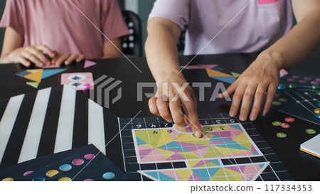 At the kitchen table, a young mother and her daughter play a board game, arranging the geometric shapes of a tangram puzzle At the kitchen table, a young mother and her daughter play a board game, arranging the geometric shapes of a tangram puzzle 117334353