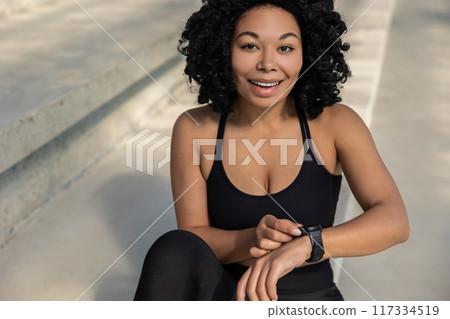 Brunette woman in sportswear sitting on the steps and looking at her smart watch 117334519