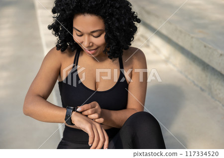 Brunette woman in sportswear sitting on the steps and looking at her smart watch 117334520