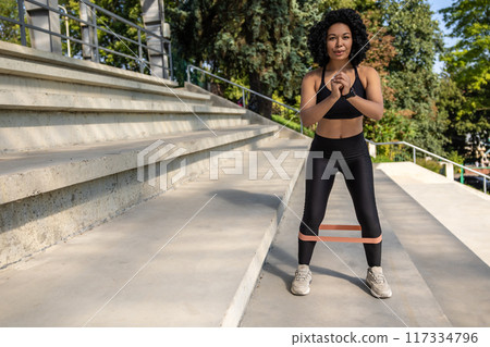 Fit woman in black sportswear exercising outdoors with a power band on her legs 117334796