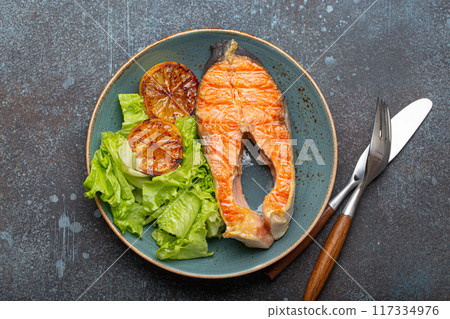 Grilled fish salmon steak and green salad with lemon on ceramic plate on rustic blue stone background top view, balanced diet or healthy nutrition meal with salmon and veggies 117334976
