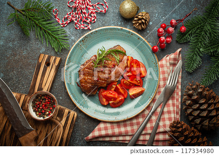 Festive Christmas dinner with roasted duck breast fillet with golden crispy skin and tomato salad, Xmas decorations on table with fir tree, cones, red berries on blue rustic concrete background. Festive Christmas dinner with roasted duck breast fillet with golden crispy skin and tomato salad, Xmas decorations on table with fir tree, cones, red berries on blue rustic concrete background. 117334980