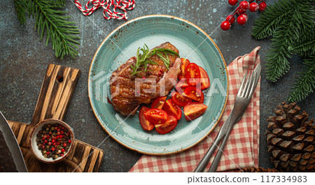 Festive Christmas dinner with roasted duck breast fillet with golden crispy skin and tomato salad, Xmas decorations on table with fir tree, cones, red berries on blue rustic concrete background. Festive Christmas dinner with roasted duck breast fillet with golden crispy skin and tomato salad, Xmas decorations on table with fir tree, cones, red berries on blue rustic concrete background. 117334983