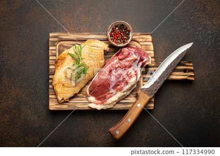 Two raw uncooked duck breast fillets with skin, seasoned with salt, pepper, rosemary top view on wooden cutting board with knife, dark brown concrete rustic background Two raw uncooked duck breast fillets with skin, seasoned with salt, pepper, rosemary top view on wooden cutting board with knife, dark brown concrete rustic background 117334990