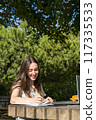 woman smiling at camera in the park with a laptop writing in a notebook  117335533