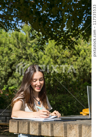 woman smiling at camera in the park with a laptop writing in a notebook  117335533