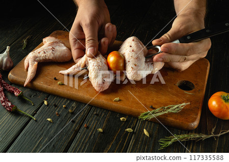 Chef hands preparing chicken wings for grill. The cook hands skewering chicken wing on fork on kitchen table. Advertisement space on black table Chef hands preparing chicken wings for grill. The cook hands skewering chicken wing on fork on kitchen table. Advertisement space on black table 117335588