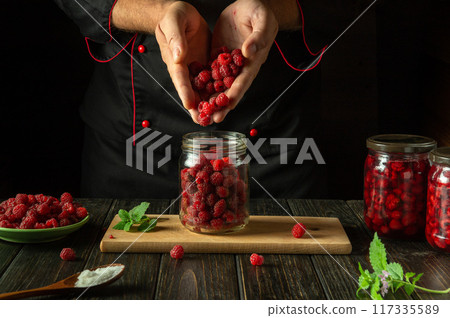 The cook prepares jam with Rubus idaeus. The concept of making sweet raspberry jam on the kitchen table. Filling the jar with ripe berries in the hands of the chef 117335589