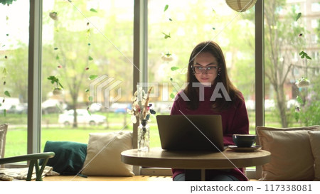 A young woman in glasses using a laptop in a cozy cafe with a panoramic window view. A young woman in glasses using a laptop in a cozy cafe with a panoramic window view. 117338081