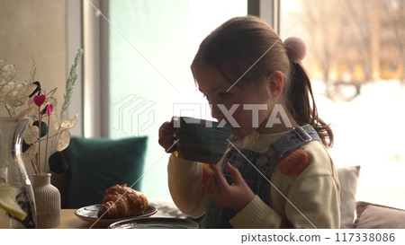 A girl drinks hot cocoa with marshmallows from a green cup against the backdrop of a cozy cafe with large panoramic windows. 117338086