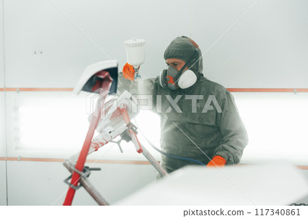 Auto mechanic worker wearing protective workwear spraying white paint on car part at workshop 117340861