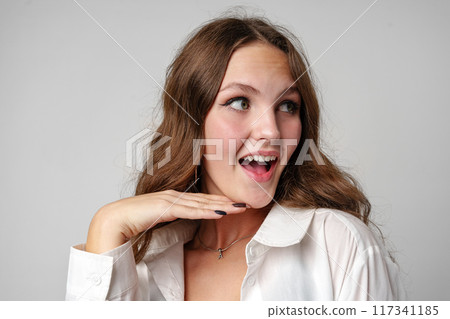 Excited Young Woman in White Blouse Reacting With Joyful Surprise Against a Grey Background Excited Young Woman in White Blouse Reacting With Joyful Surprise Against a Grey Background 117341185