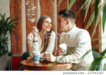 Smiling Young Couple Enjoying Coffee and Conversation in a Cozy Cafe Setting Smiling Young Couple Enjoying Coffee and Conversation in a Cozy Cafe Setting 117341287