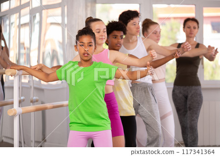 Dark-skinned girl practicing demi plie at barre with group 117341364