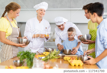 Young guy and adult woman cook teaches group of children to cook Young guy and adult woman cook teaches group of children to cook 117341700
