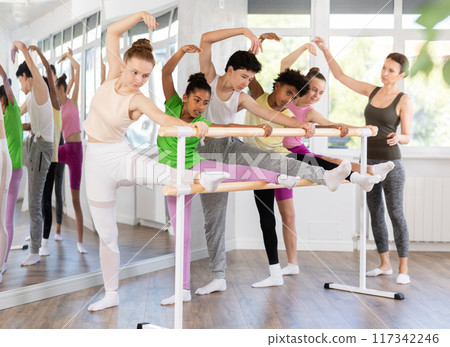 Teen girl performing stretching exercises at ballet barre in group class 117342246