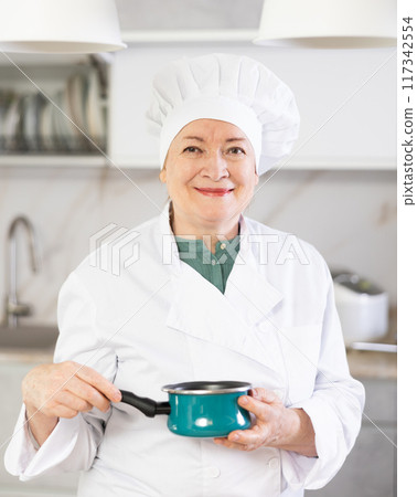 Elderly woman in chef hat and white uniform stands in front of table and holds stewpot 117342554