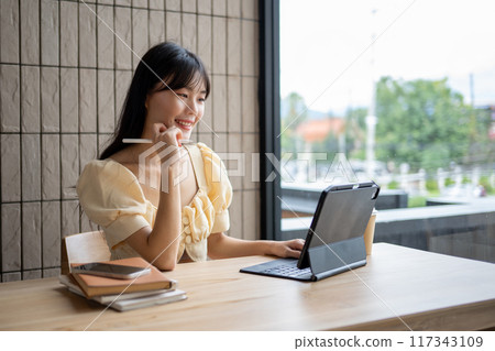 A beautiful Asian woman working on her digital tablet in a modern coffee shop, working remotely. 117343109