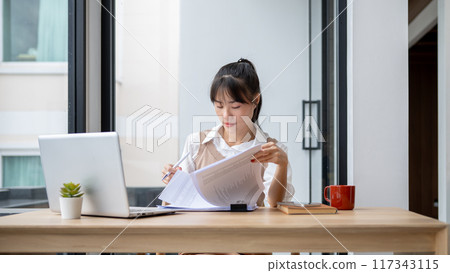 A focused Asian businesswoman is reviewing business report at her desk, working in her office. 117343115
