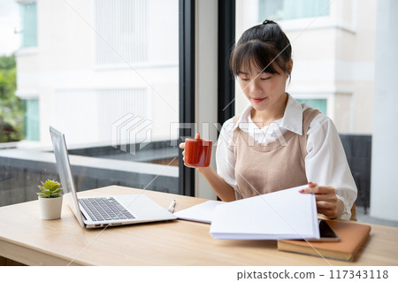 A beautiful Asian businesswoman having a morning coffee while reviewing business report at her desk. 117343118