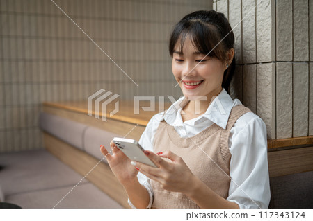 A happy Asian woman is enjoying chatting with her friends on her smartphone while relaxing indoors. 117343124