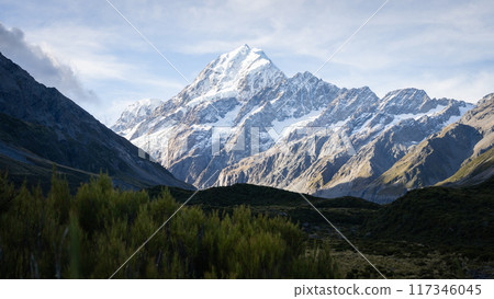 Massive snowy mountain with glacier catching the last light and towering above shaded alpine valley 117346045