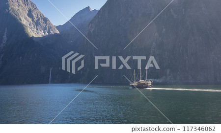 Boat sailing through fjord with steep rocky walls and waterfall in distance, Fiordland, New Zealand 117346062