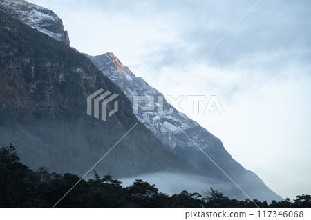 Rocky mountain walls catching first sunlight during hazy autumn morning in Fiordland, New Zealand 117346068