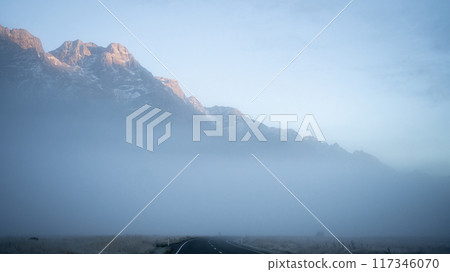 Empty highway leading through the foggy valley with mountains in background catching early sunlight 117346070