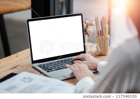 A woman typing on a laptop with a blank screen at a wooden desk in a modern office setting, illuminated by natural light. 117346330
