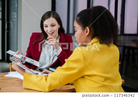 Two professional women engaged in a business meeting, reviewing documents in a modern office environment. Two professional women engaged in a business meeting, reviewing documents in a modern office environment. 117346342