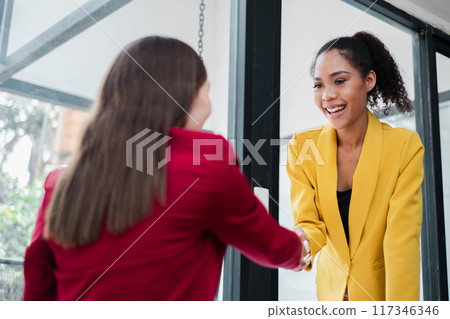 Two women in professional attire shaking hands in a modern office setting, symbolizing agreement, partnership, and collaboration. 117346346