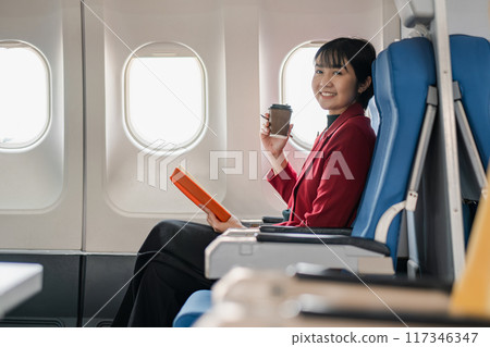 A businesswoman in a red blazer enjoys coffee and reads a book while seated on an airplane, showcasing in-flight comfort and productivity. 117346347