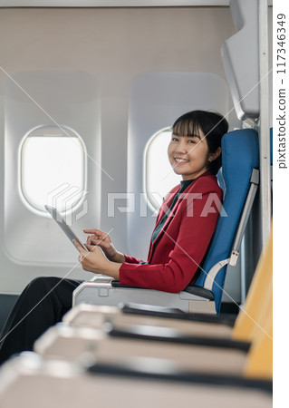 Smiling businesswoman in red blazer using a tablet while seated in an airplane's economy class. 117346349