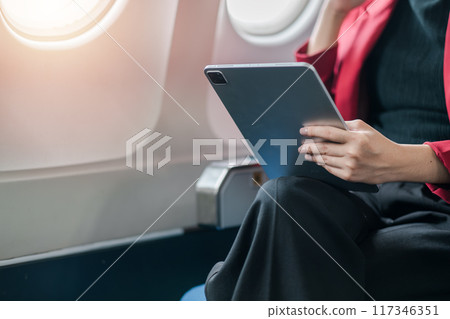 A businesswoman in a red blazer using a tablet while seated on an airplane, showcasing modern travel and technology. 117346351