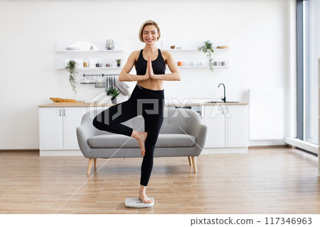 Woman practicing yoga at home in tree pose in living room Woman practicing yoga at home in tree pose in living room 117346963