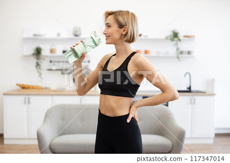 Woman in sporty outfit drinking water in modern kitchen 117347014