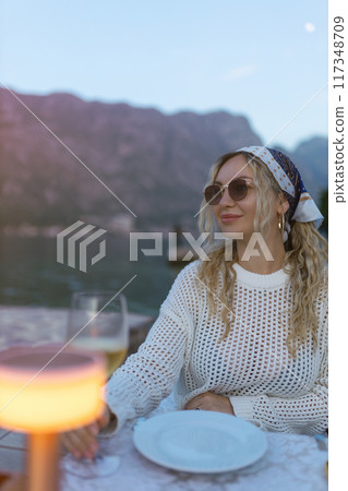 Young woman having dinner in a hotel restaurant during sunset near the sea and mountains, close-up. A girl enjoys the sunset with a glass of wine. Vacation and travel concept 117348709