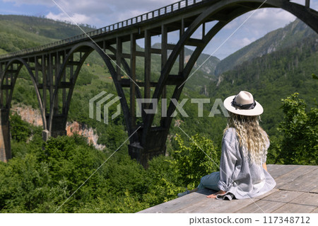 A girl in a hat stands against the backdrop of a beautiful arched bridge over the Tara River, a popular tourist attraction in Montenegro. 117348712