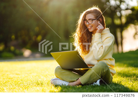 Young woman with wireless headphones working online or studying sitting on grass at park. Blogging Young woman with wireless headphones working online or studying sitting on grass at park. Blogging 117349876