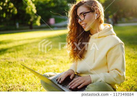 Young woman with wireless headphones working online or studying sitting on grass at park. Blogging Young woman with wireless headphones working online or studying sitting on grass at park. Blogging 117349884