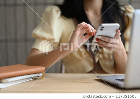 Close up of young woman typing text message on mobile phone while sitting at office desk 117351503