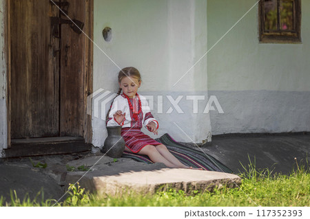 A young girl dressed in traditional attire stands by an old rustic door in warm summer sunlight A young girl dressed in traditional attire stands by an old rustic door in warm summer sunlight 117352393
