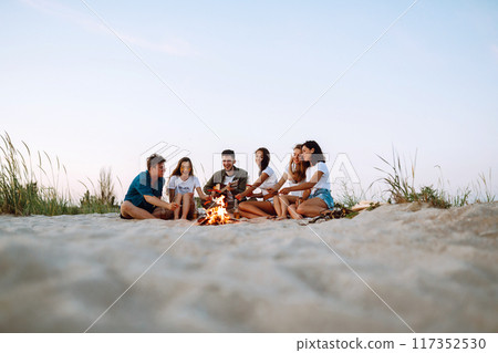 Group of young friends sitting on beach and fry sausages. One man is playing guitar. Camping time. 117352530