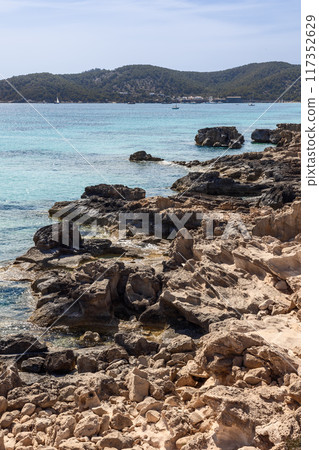 Ibiza rugged coast with clear turquoise waters, contrasting shoreline, distant hills 117352629